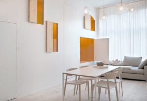 A modern, minimalist living room with a white table and chairs, light wooden flooring, white curtains, and orange accent piec