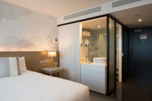 Modern minimalist hotel room with a white bed, glass door leading to a bathroom, and a wooden headboard.
