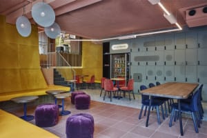 A modern cafe with yellow seating, blue chairs, purple poufs, and a vending machine.