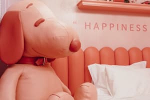 A large, pink, stuffed dog toy wearing a red bow tie lying on a bed with white pillows.