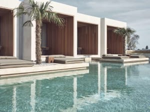 Concrete poolside lounge area with wooden shades, palm trees, and clear blue water.