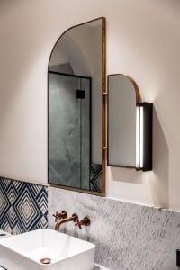 Modern bathroom featuring metallic brass accents and wall-mounted light fixture with marble tiled backdrop.
