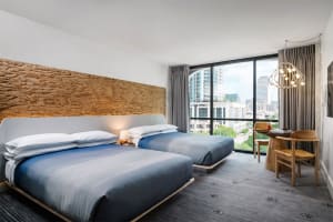 A modern hotel room with two beds, a desk, and a large window providing a view of the city.