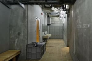 A modern public bathroom with concrete walls, a steel sink, and a wooden bench.