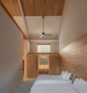 A minimalist bedroom with wooden walls and beds, featuring a ceiling fan, and a bathroom in the background.