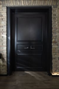 A modern, black wooden door with a panel design, set against a textured brick wall.