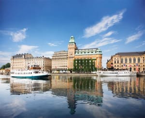 A cityscape featuring stone and glass buildings reflected in calm water.