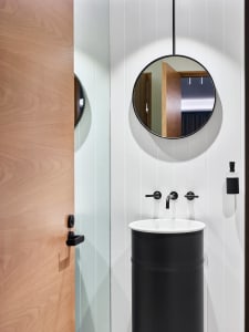 Modern bathroom interior featuring a wooden door, a black pedestal sink with a round mirror above, and a black faucet.