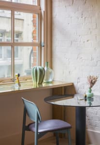 A minimalist window-side table with a wooden frame, glass tabletop, and a green plant and vase on display.