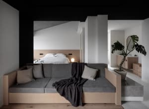 A minimalist living room featuring a floating sofa with gray cushions and a black throw blanket, a concrete headboard bed wit