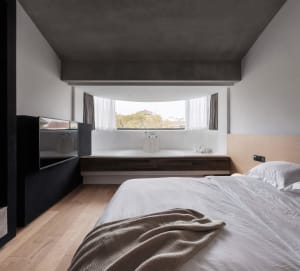 A modern minimalist bedroom with concrete walls, wood flooring, and a large window.