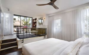 A modern bedroom featuring a glass wall with a view of the outdoors, a white bed with gold accents, and a balcony.