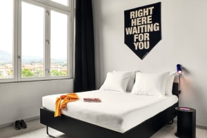 A minimalist bedroom featuring a white bed with a black headboard and a black table lamp, with a banner reading 'Right Here W