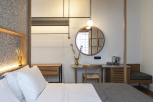 A modern minimalist bedroom with wooden furniture and white bedding, featuring a round mirror, a small table, and a comfortab