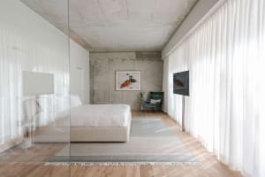 A modern minimalist bedroom with a concrete wall, white bed, and white curtains.