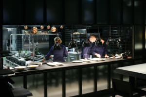 Three chefs preparing food in a modern, industrial-style restaurant kitchen with metal, glass, and stainless steel elements.