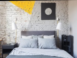Modern industrial bedroom with gray upholstered bed and striped bedding against a weathered brick wall with geometric mural