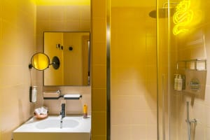 A minimalist bathroom with yellow tiles, featuring a white sink, a mirror, and a neon light fixture in the shape of a snake.