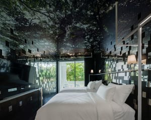 A modern bedroom featuring black mirrored tiles, a window with a view of trees, a twin bed with white bedding, and a flat-scr