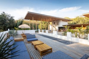 A modern residential patio featuring wooden furniture, metal railings, and fabric cushions under a large wooden canopy.