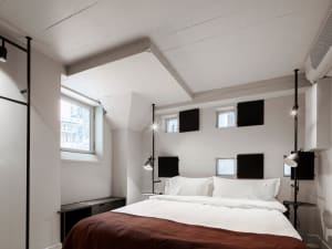 A minimalist bedroom featuring a bed with white bedding and a red blanket, minimalistic decor, and natural light from a windo