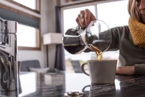 A cozy kitchen with a stone countertop and a woman pouring coffee into a mug from a glass carafe.