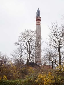 A tall, industrial concrete smokestack with red and gray stripes, surrounded by bare trees and autumn foliage.