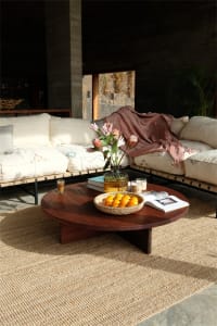 A modern living room featuring a round wooden table with a bowl of oranges, a vase of flowers, and various other items.