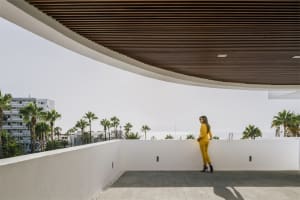 A woman in a yellow suit standing on a modern balcony with steel and white concrete elements, overlooking a coastal view with