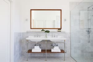 A modern bathroom with white marble tile walls, floor, and two white sinks with chrome faucets and a wooden shelf below. A gr