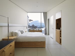 A modern minimalist bedroom features a large bed with white bedding, a glass window, and wooden furniture with a view of a mo
