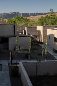 Minimalist courtyard with concrete walls and stone paths, surrounded by greenery and hills in the background.