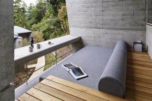 A minimalist living room with a grey sofa, a wooden bench, a concrete wall, and a view of greenery outside.