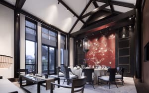 A modern restaurant dining room with a dark wooden ceiling and walls, natural light from large windows, and a round dining ta