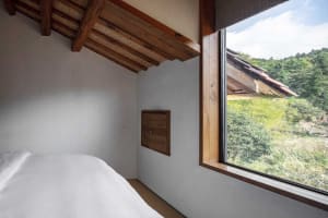 A minimalist bedroom with a white bed and a large window overlooking greenery.