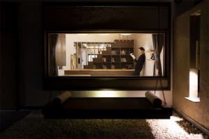 A man reads a book on a floating platform in an industrial-style living space with a bookshelf and wooden elements.