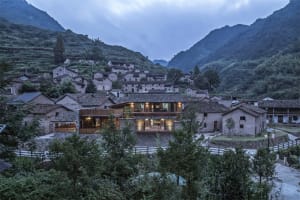 A traditional stone house with a clay-tiled roof nestled among lush green mountains.