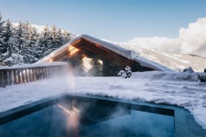 A modern industrial swimming pool with mist rising from the water, surrounded by snow-covered trees and a building with a woo