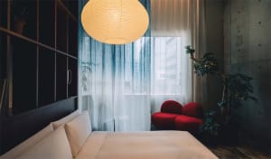 Modern minimalist hotel room with white textiles, red furniture, and a large window with blue curtains.
