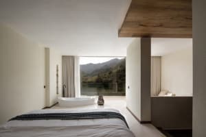 A modern bedroom with a concrete floor, a white bed with a black blanket, and a freestanding bathtub overlooking a scenic mou