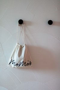 A white fabric bag with 'Momos' written in black cursive letters, hanging on a wall with a wave-pattern design.