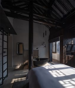 Interior of a minimalist bedroom with concrete walls and ceiling beams, featuring a black bathtub and a bed.