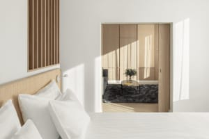 Modern, minimalist bedroom with white bedding, wooden furniture, and light streaming through the window.