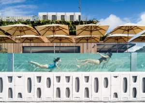 People swimming underwater in a transparent glass rooftop pool with umbrellas and plants in the background.