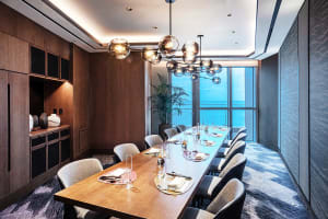 A modern restaurant dining room featuring a wooden table and chairs, chandelier, and ocean view.