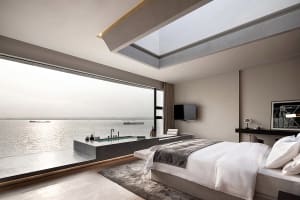 A modern minimalist bedroom featuring a concrete bathtub with a view of the water and a large window allowing natural light t
