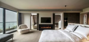 Modern minimalist bedroom featuring a large bed with white bedding, a glass fireplace, and large windows with glass walls.