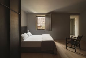 A minimalist bedroom with a bed, chair, and small window overlooking a tiled rooftop.