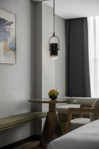 A modern minimalist bedroom featuring a wooden table with a small plant, a hanging metal lantern, and a bed with a white blan