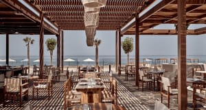 An outdoor restaurant setting with wooden tables, chairs, and a pergola structure overlooking the sea.